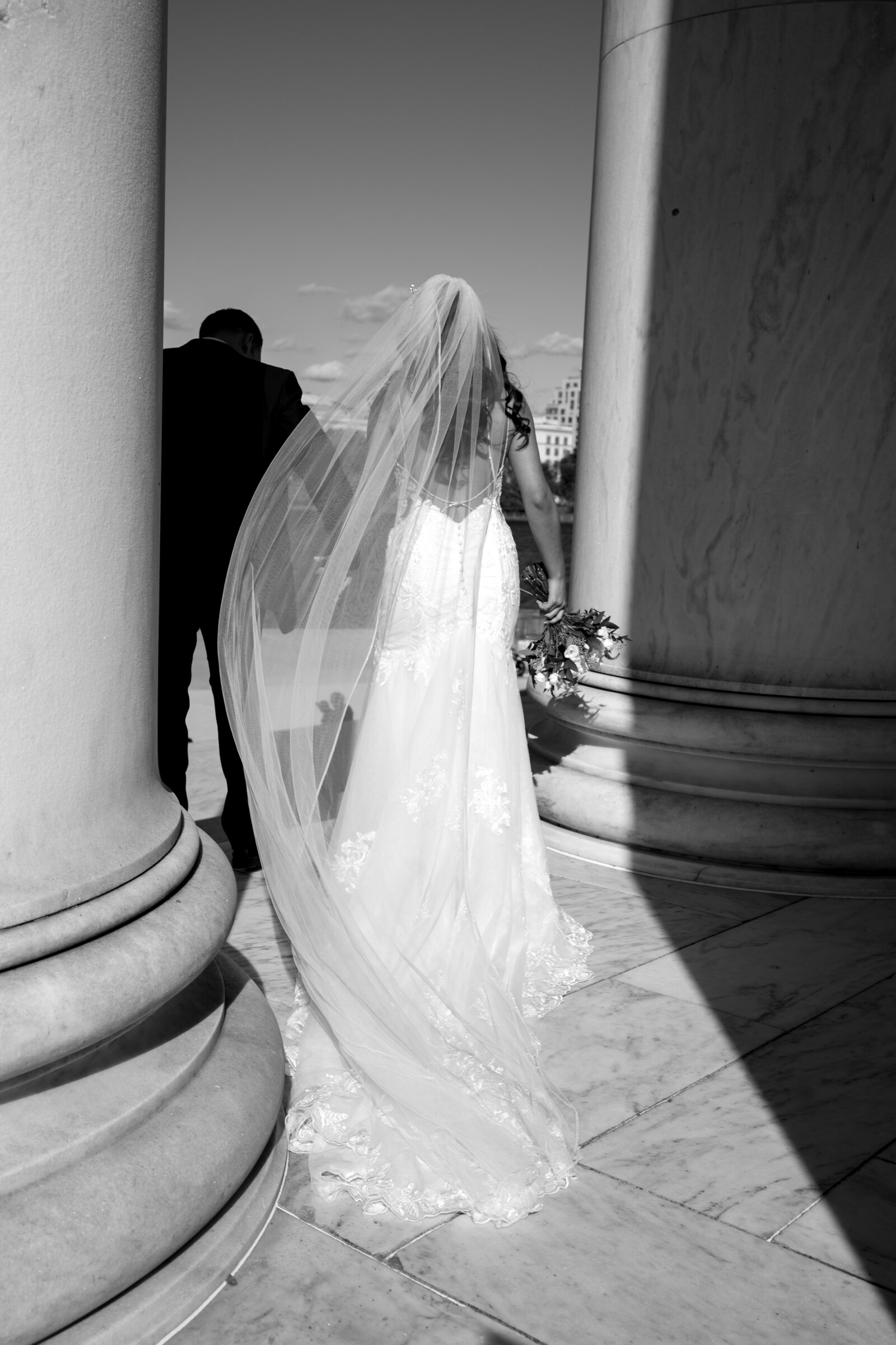 A cathedral veil caught in the wind between two marble columns, her groom a step ahead in shadow.